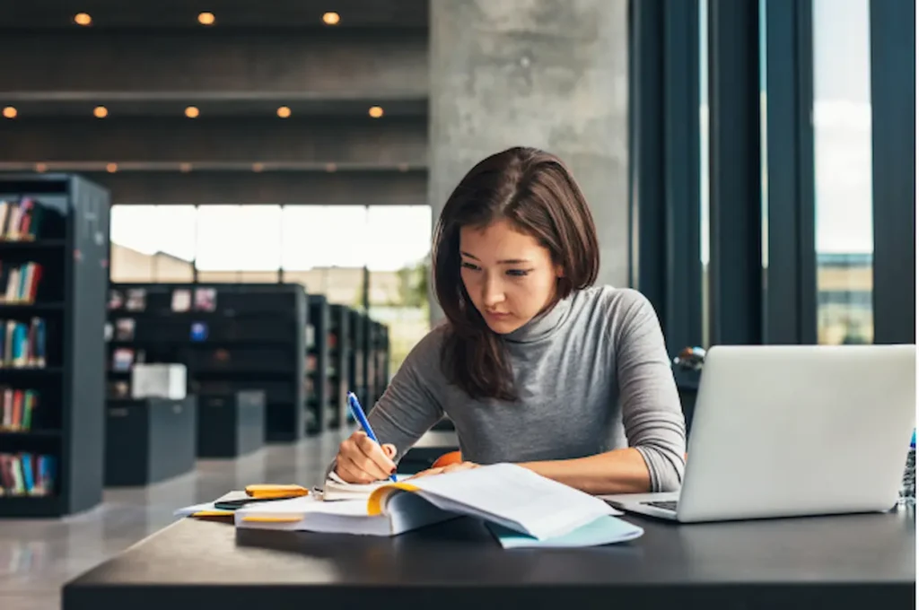 Studentessa concentrata che prende appunti in biblioteca, con un laptop e libri di testo aperti davanti a sé.